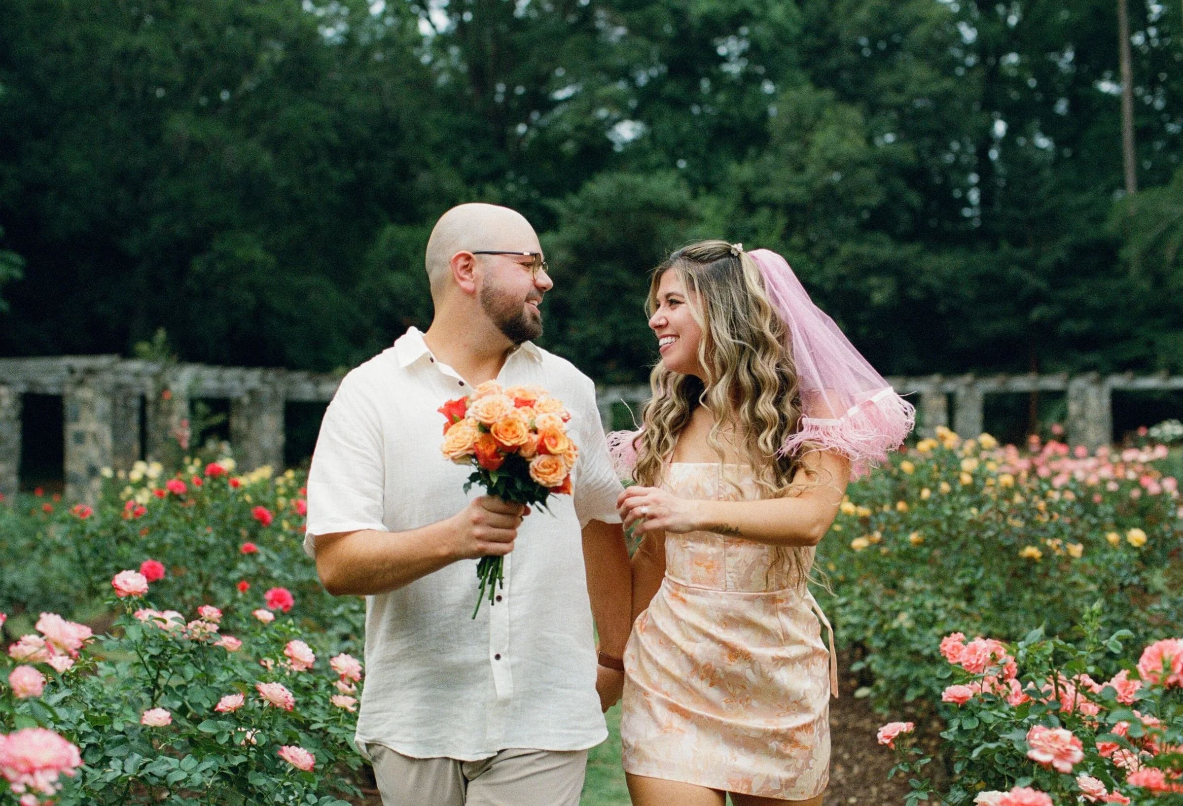 Nicole and Ian walking through the rose garden, Nicole in a pink veil holding a bouquet of peach roses