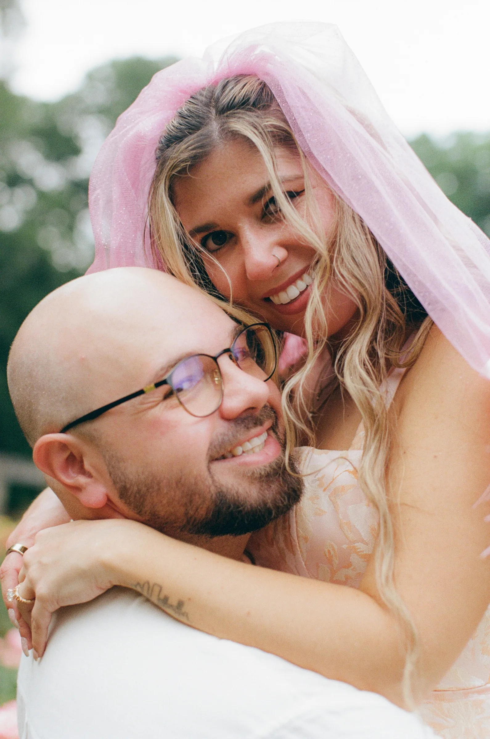 Nicole hugging Ian from behind, both smiling, her pink veil over them