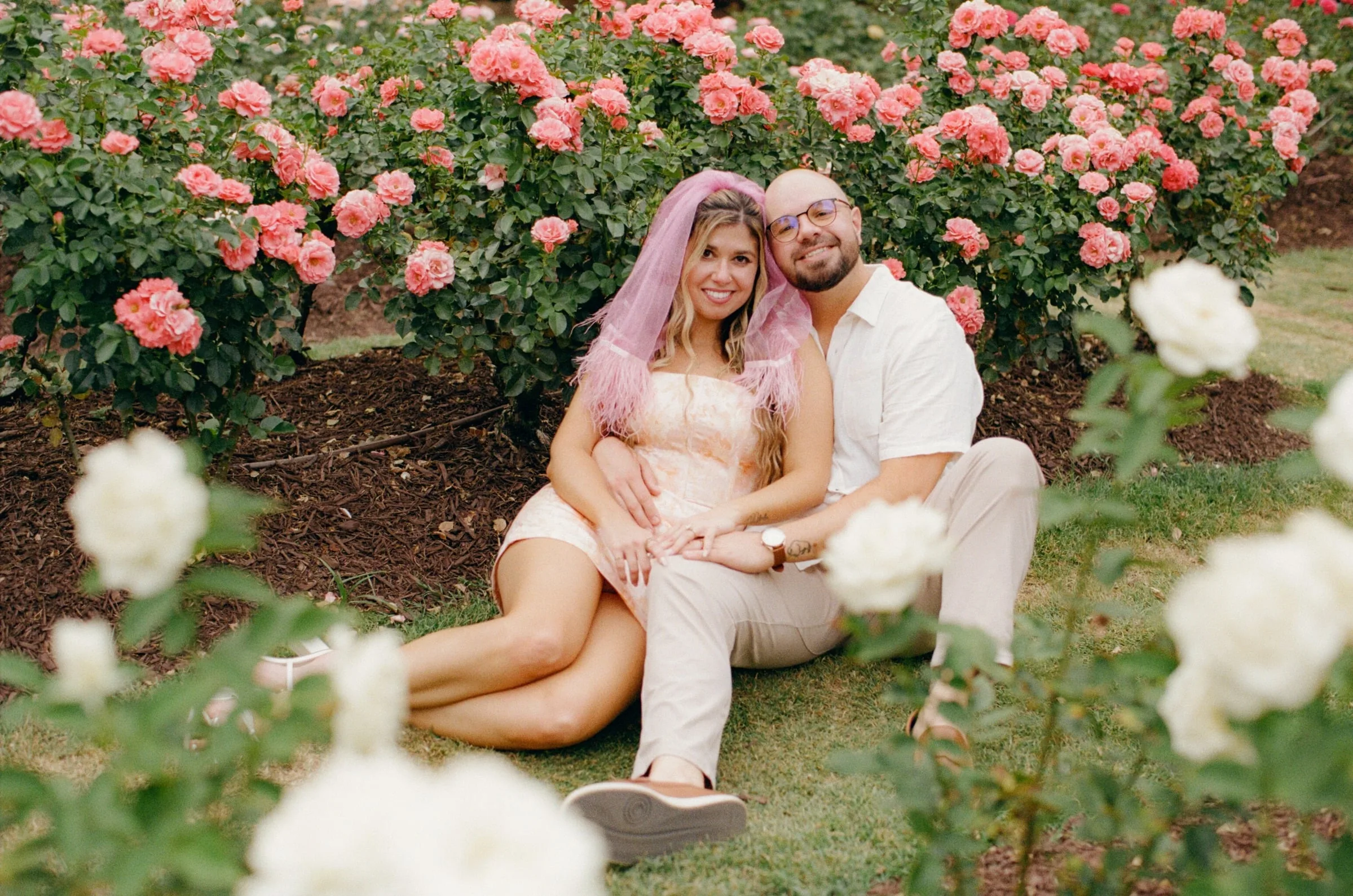 Nicole and Ian sitting on the grass in front of a bed of pink roses